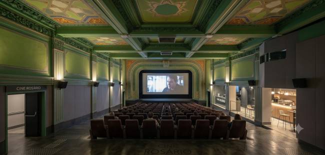 Interior de um cinema com poltronas marrons vazias, exceto por algumas pessoas sentadas. Uma tela grande exibe um filme com um homem. As paredes e o teto s&atilde;o verdes com detalhes ornamentados e ilumina&ccedil;&atilde;o indireta. &Agrave; direita, h&aacute; uma &aacute;rea de caf&eacute; com balc&atilde;o e bancos. &Agrave; esquerda, uma porta com a inscri&ccedil;&atilde;o CINE ROS&Aacute;RIO